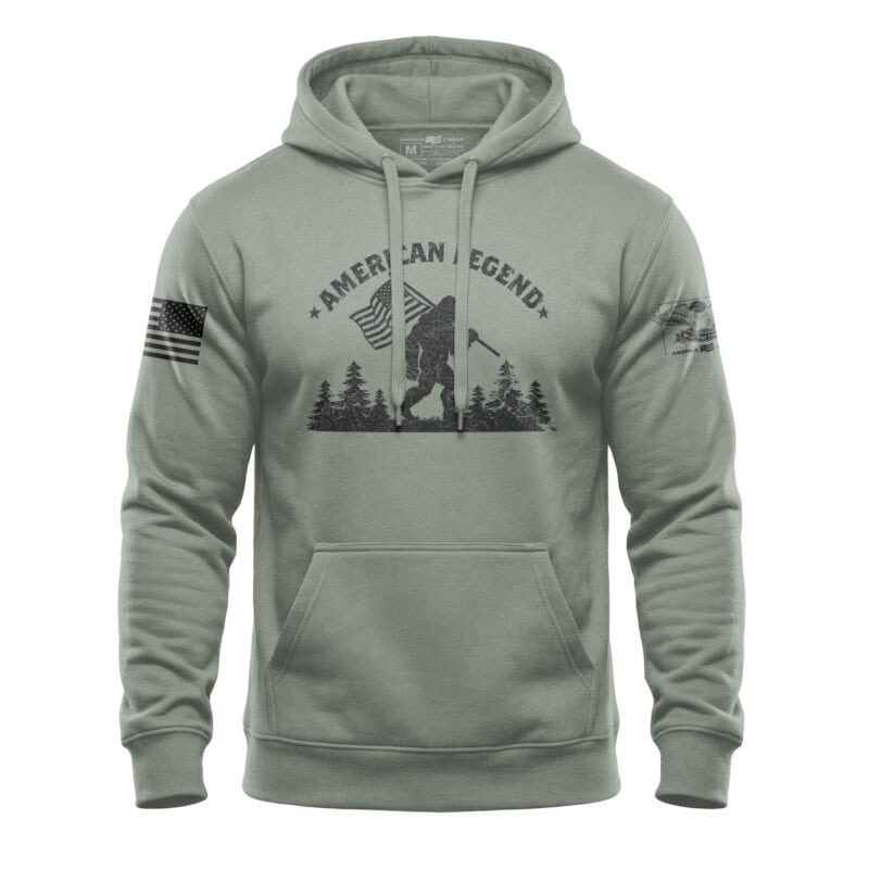 Patriotic Bigfoot hoodie with American flag and forest design in sage green. Perfect for outdoor ent.